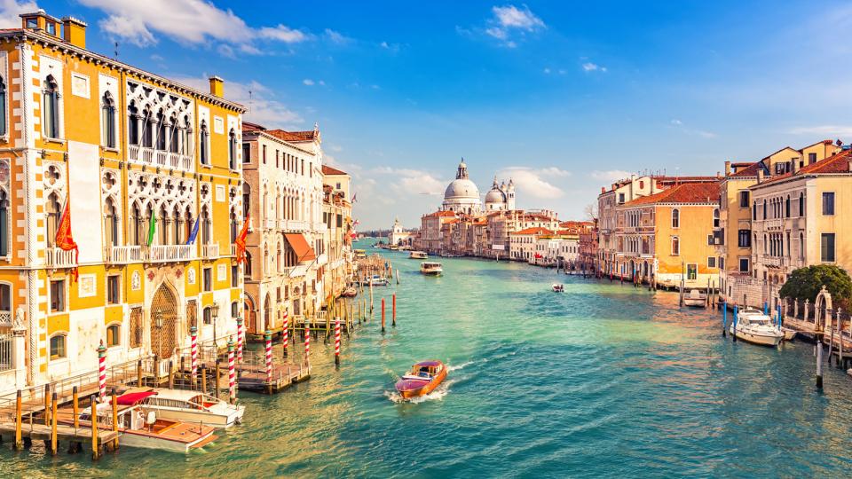Ville de Venise. Le paysage est ensoleillé, la ville très colorée (particulièrement dans des tons jaunes et orangés). Un bras d'eau divise la Ville.